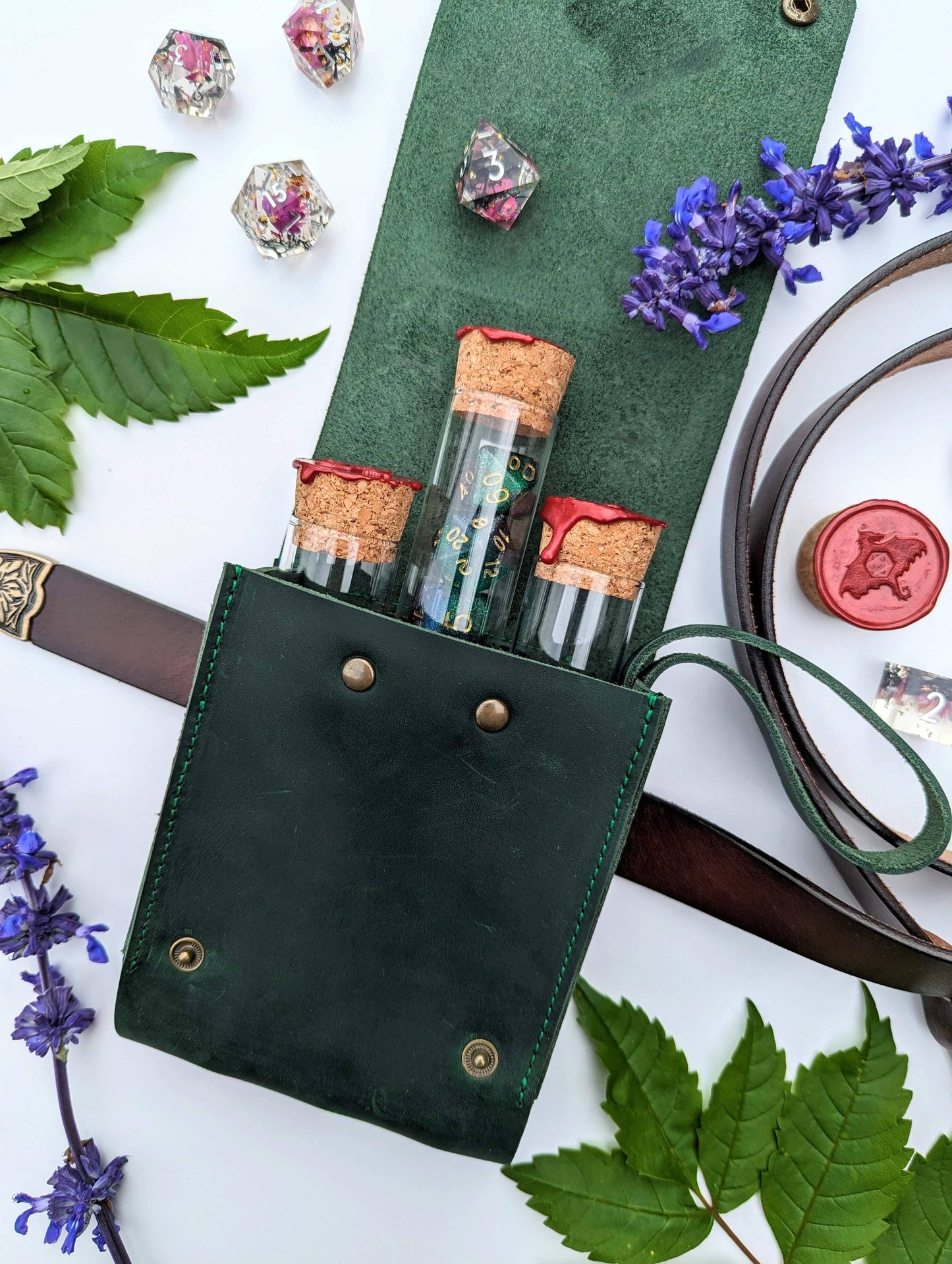 A green genuine leather pouch with a built-in belt loop and snap closures, designed to hold glass vials with dice inside, displayed with flowers and leaves around it.